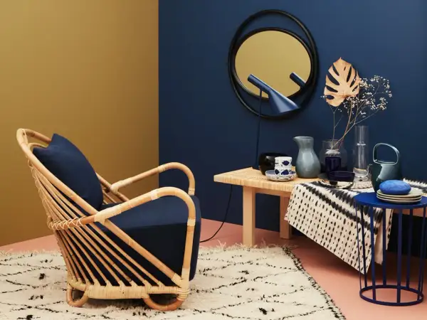 Ein Rattansessel mit dunkelblauem Kissen steht auf einem flauschigen Teppich. Daneben ein Tisch mit Deko. Welche Farbe passt zu Gold? Dunkelblau und Senfgelb harmonieren perfekt.