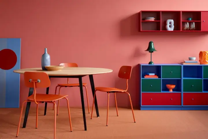 Ein Essbereich mit roten Stühlen und einem runden Holztisch. Die Wand ist korallenrot, und ein blaues Regal mit roten Akzenten steht daneben. Welche Farbe passt zu Rot? Blau und Grün sind tolle Kontraste!
